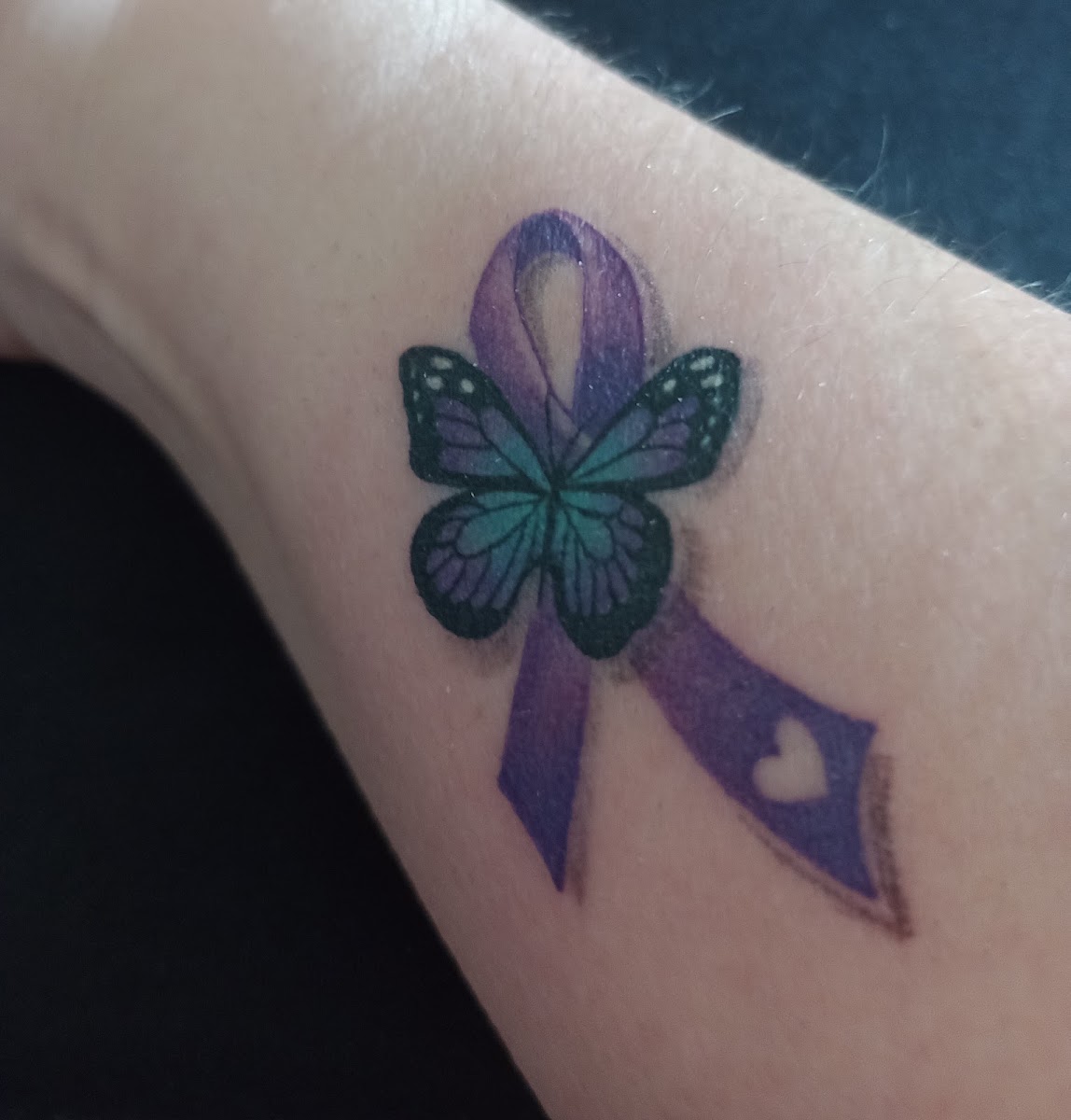 Tatuaje cicatrizado de cinta y mariposa en tonos violeta y verde en el brazo, trabajo real de Crabink Tattoo